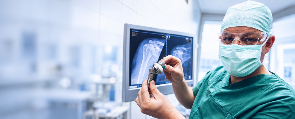 Dr. med. Tobias Merkle in OP-Kleidung mit einer Schulterprothese vor einer Röntgenaufnahme der Schulter